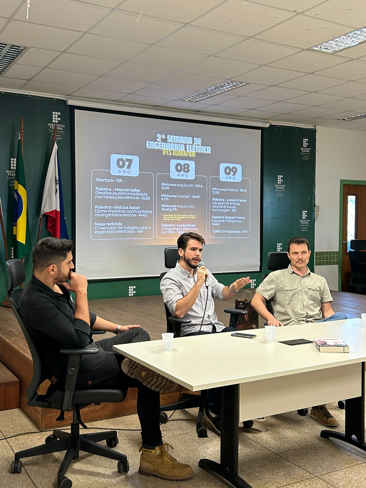 Em uma mesa pr&oacute;xima ao palco, est&atilde;o tr&ecirc;s palestrantes. Um deles fala ao microfone. Na proje&ccedil;&atilde;o de tela, ao fundo, est&aacute; exibida a programa&ccedil;&atilde;o do evento.
